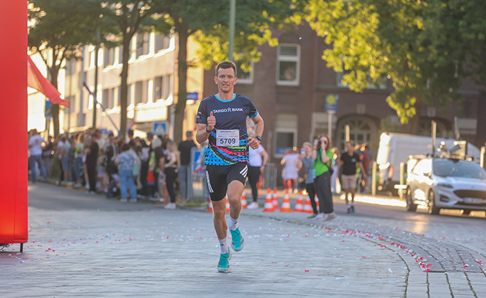 Ein Läufer mit der Nummer 5709 läuft auf einer Straße, umgeben von Zuschauern. Er trägt ein schwarzes Trikot mit blauen und weißen Mustern, kurze schwarze Shorts und grüne Laufschuhe. Im Hintergrund sind Menschenmengen zu sehen, die ihn anfeuern, sowie eine rote Bogenstruktur und Autos. Die Straße ist mit Konfetti bedeckt.