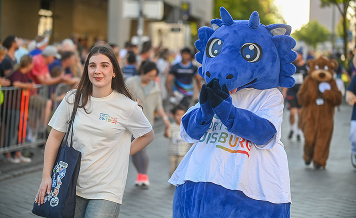 Eine junge Frau mit dunklen Haaren trägt ein helles T-Shirt und eine Tasche. Neben ihr steht ein blauer Drachenkostümträger mit einem weißen T-Shirt, das 'Duisburg' zeigt. Im Hintergrund sind viele Menschen auf einer belebten Straße zu sehen.