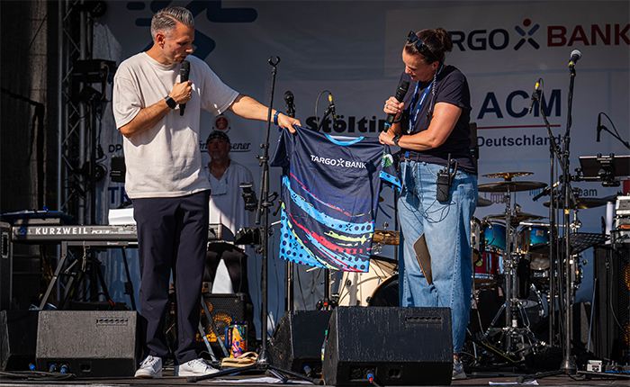 Eine Veranstaltung auf einer Bühne, wo ein Mann und eine Frau miteinander sprechen. Der Mann hält ein Mikrofon und zeigt auf ein T-Shirt mit dem Aufdruck 'TARGO X BANK', das die Frau in der Hand hält. Im Hintergrund sind Musiker und Instrumente auf der Bühne sichtbar. Es ist sonnig und die Atmosphäre ist festlich.