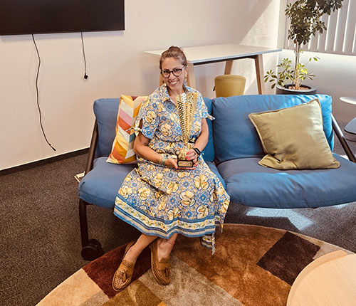 Eine Frau sitzt auf einem blauen Sofa in einem modernen Raum. Sie trägt ein hellblaues Kleid mit gelben Blumenmotiven und eine lange Kette. In ihrer Hand hält sie einen Preis. Im Hintergrund sind Pflanzen und ein Tisch sichtbar.