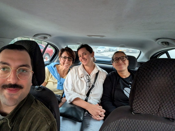 Eine Gruppe von vier Personen sitzt im hinteren Teil eines Autos. Der Mann links hat einen Schnurrbart und trägt eine Brille. Die Frauen neben ihm lächeln. Eine Frau trägt ein blaues Oberteil, eine andere ein weißes Hemd. Die dritte Frau hat ein schwarzes T-Shirt an. Im Hintergrund sind andere Autos sichtbar.