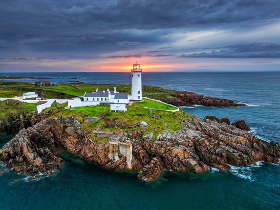 Ein Leuchtturm steht auf einer Felseninsel, umgeben von türkisblauem Wasser. Im Hintergrund sind dunkle Wolken und ein Sonnenuntergang sichtbar. Die Gebäude rund um den Leuchtturm sind weiß und gut erhalten. Grüne Wiesen umgeben die Struktur, während die Felsen ins Wasser ragen.