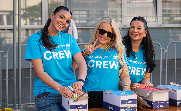 Drei Frauen in blauen T-Shirts mit der Aufschrift "CREW" stehen an einem Tisch. Sie lächeln und halten eine Handvoll Snacks, während sie in Kisten mit Snacks arbeiten. Im Hintergrund sind Gitter und eine graue Wand sichtbar.