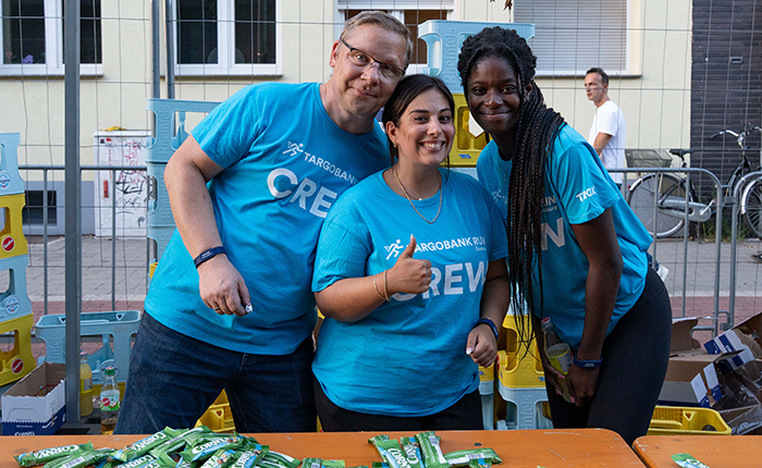 Drei Personen stehen an einem Tisch mit Snacks. Sie tragen blaue T-Shirts mit dem Aufdruck 'CREW' und dem Logo der TARGOBANK. Die Atmosphäre ist freundlich.