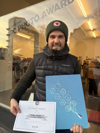 Ein Mann steht vor einem Fenster, das das Schild "SPC PHOTO AWARD" zeigt. Er trägt eine grüne Mütze und eine schwarze Weste. In seiner rechten Hand hält er ein Zertifikat mit der Aufschrift "SPC PHOTO AWARDS" und in seiner linken Hand ein blaues Poster mit weißen Kreisen. Im Hintergrund sind mehrere Personen zu sehen.