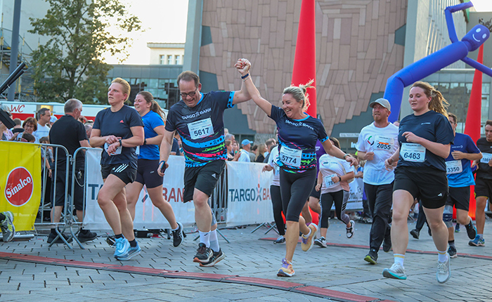 Eine Gruppe von Läufern nimmt an einem Rennen teil. Zwei Personen, die Hände haltend und in Sportkleidung, überqueren die Ziellinie. Die Läufer tragen Nummern auf ihren Oberteilen. Im Hintergrund sind Zuschauer und Werbebanner sichtbar, sowie ein aufblasbares Luftfigur. Es ist ein sonniger Tag.