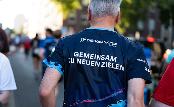 Ein Mann mit grauen Haaren trägt ein T-Shirt mit der Aufschrift "GEMEINSAM ZU NEUEN ZIELEN" und dem Logo der TARGOBANK RUN Duisburg. Er steht in einer Gruppe von Menschen auf einer Straße, während es hell ist.