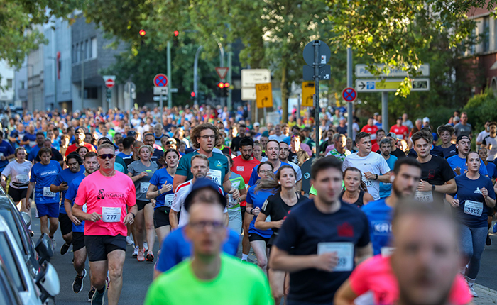 Eine große Gruppe von Läufern läuft auf einer Straße, umgeben von Bäumen und städtischen Gebäuden. Viele tragen Sportkleidung in verschiedenen Farben, darunter ein Mann in einem pinken Shirt mit der Nummer 777. Die Szene zeigt ein aktives und lebhaftes Rennen mit zahlreichen Teilnehmern, die sich in Richtung der Kamera bewegen.