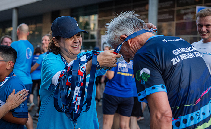 Eine Frau in einem blauen Shirt und einer Kappe hängt einer älteren männlichen Person eine Medaille um. Der Mann trägt ein sportliches Shirt mit dem Text 'GEMEINSAM ZU NEUEN ZIELEN'. Im Hintergrund sind mehrere Menschen in blauen Shirts zu sehen, die den Moment beobachten. Die Szene wirkt festlich und feierlich.