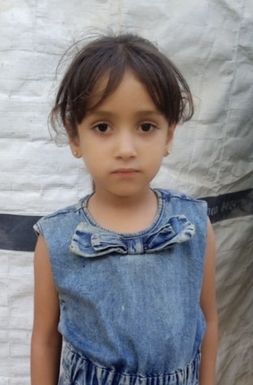 Ein kleines Mädchen mit dunklen, langen Haaren steht vor einer weißen Wand. Sie trägt ein ärmelloses, blaugraues Kleid mit einer Schleife am Kragen. Ihr Gesichtsausdruck ist neutral und ernst.