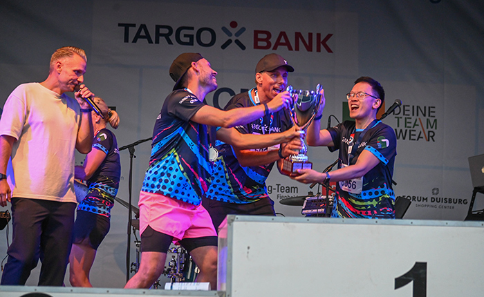 Eine Gruppe von vier Männern auf einer Bühne, die den ersten Platz in einem Wettbewerb feiern. Sie tragen bunte Sporttrikots mit dem Logo der TARGOBANK. Ein Mann hält einen Pokal, während die anderen jubeln. Im Hintergrund steht eine Person mit einem Mikrofon. Es gibt eine große Banner mit dem Schriftzug TARGOBANK und andere Sponsorenlogos.