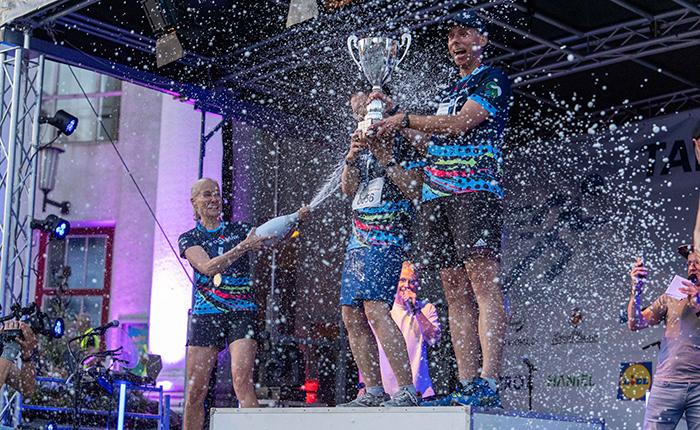 Drei Personen stehen auf einer Siegesbühne und halten einen Pokal in die Höhe. Sie sind mit sportlichen T-Shirts in bunten Farben gekleidet. Um sie herum spritzt Wasser oder Champagner, während eine jubelnde Menge im Hintergrund zu sehen ist. Die Atmosphäre ist festlich und feierlich.