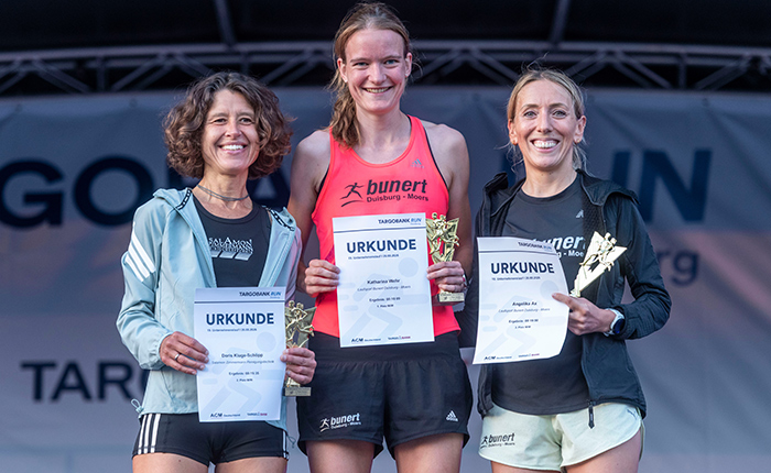 Drei Frauen stehen auf der Bühne mit Urkunden und Trophäen in der Hand. Die Frau in der Mitte trägt ein rotes Sporttop mit dem Logo 'bunert' und lächelt. Die beiden anderen Frauen sind links und rechts von ihr, eine in einem grauen Oberteil und die andere in einem schwarzen. Alle drei zeigen stolz ihre Urkunden, auf denen 'URKUNDE' steht.