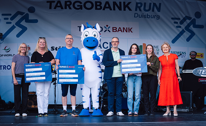 Eine Gruppe von sechs Personen steht auf einer Bühne beim TARGOBANK RUN in Duisburg. In der Mitte steht ein Maskottchen, ein weißes Pferd mit blauen Akzenten. Alle Personen halten große Plakate mit dem Logo der TARGOBANK. Die Frauen tragen verschiedene Outfits, darunter ein rotes Kleid, während ein Mann in einem blauen T-Shirt zu sehen ist. Im Hintergrund ist eine Bühnenkonstruktion mit einem großen Banner, das den Schriftzug "TARGOBANK RUN" zeigt.