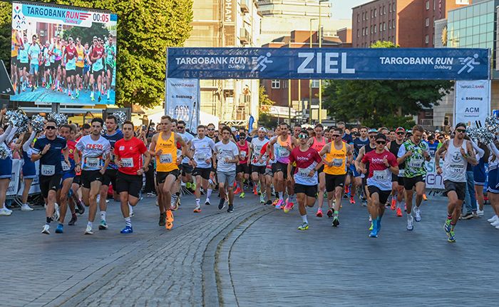 Eine große Gruppe von Läufern startet beim TARGOBANK RUN. Im Vordergrund sieht man viele Teilnehmer in verschiedenen Trikots, darunter rote, gelbe und weiße Farben. Sie laufen unter einem Banner mit der Aufschrift 'ZIEL'. Im Hintergrund ist eine große digitale Anzeige zu sehen, die die Zahl 00:05 zeigt. Die Umgebung ist urban mit Gebäuden und Bäumen.