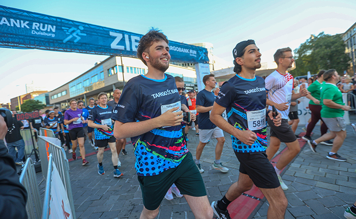 Zwei junge Männer laufen während des TARGOBANK RUN in Duisburg. Sie tragen bunte Laufshirts und lächeln. Im Hintergrund sind weitere Teilnehmer und eine große Zielbanner sichtbar.