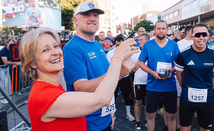 Eine Gruppe von Menschen steht an der Startlinie eines Laufs. Eine Frau in einem roten Oberteil lächelt und hält ein Smartphone, während ein Mann in einem blauen T-Shirt neben ihr steht. Im Hintergrund sind weitere Teilnehmer zu sehen, die ebenfalls in sportlicher Kleidung gekleidet sind. Ein großes Display zeigt eine Veranstaltung im Hintergrund.