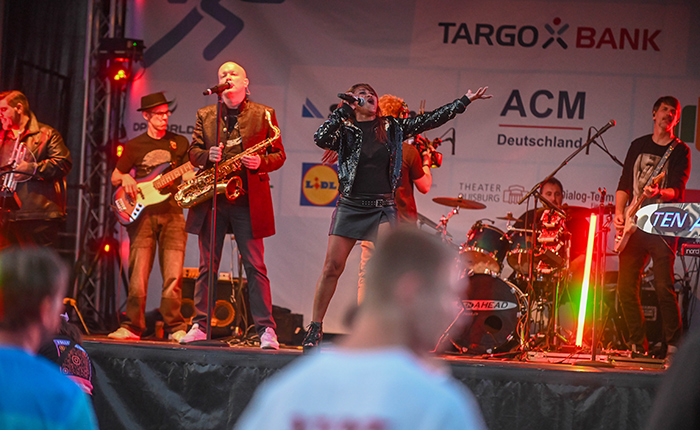 Eine Band tritt auf einer Bühne auf, während das Publikum zuschaut. Die Sängerin in einem glänzenden Outfit singt, während ein Mann mit einem Saxophon neben ihr steht. Weitere Bandmitglieder spielen Gitarre und Schlagzeug. Im Hintergrund sind Werbebanner sichtbar, darunter TARGO BANK und ACM Deutschland.