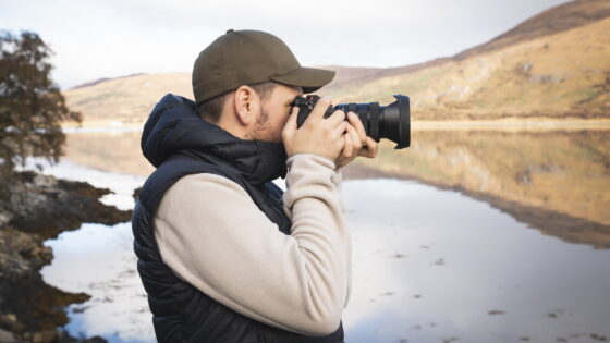 Ein Mann steht am Wasser und hält eine Kamera, um ein Foto zu machen. Er trägt eine schwarze Weste über einem hellen Pullover und eine grüne Mütze. Im Hintergrund sind Berge und ein ruhiges Gewässer zu sehen.