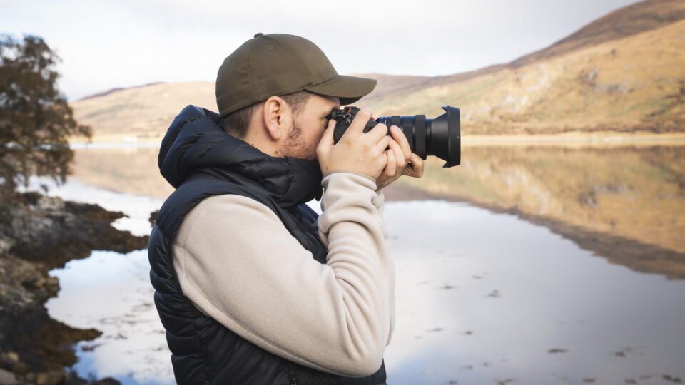 Ein Mann steht am Wasser und hält eine Kamera, um ein Foto zu machen. Er trägt eine schwarze Weste über einem hellen Pullover und eine grüne Mütze. Im Hintergrund sind Berge und ein ruhiges Gewässer zu sehen.