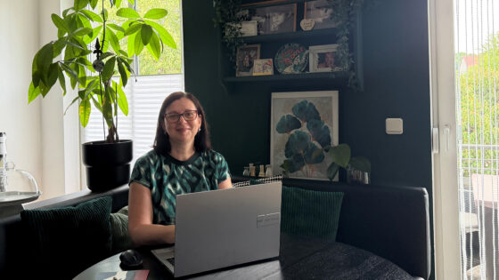 Eine Frau mit Brille sitzt an einem Tisch mit einem Laptop. Im Hintergrund sind Pflanzen und Bilderrahmen sichtbar. Die Wand ist dunkelgrün.