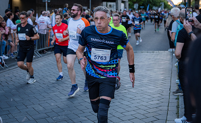 Eine Gruppe von Läufern nimmt an einem Straßenlauf teil. Ein Mann mit grauen Haaren und einer Brille trägt ein buntes T-Shirt mit dem Logo von TARGOBANK und hat die Startnummer 5763. Er läuft schnell, während andere Teilnehmer in verschiedenen Laufoutfits hinter ihm sind. Die Umgebung ist urban, mit Zuschauern auf der linken Seite, die an der Strecke stehen.