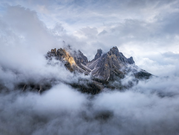 Eine beeindruckende Berglandschaft mit schroffen Gipfeln, die von Wolken umhüllt sind. Die Berge reflektieren ein sanftes Licht, während Nebel sanft über die bewaldeten Hänge zieht.
