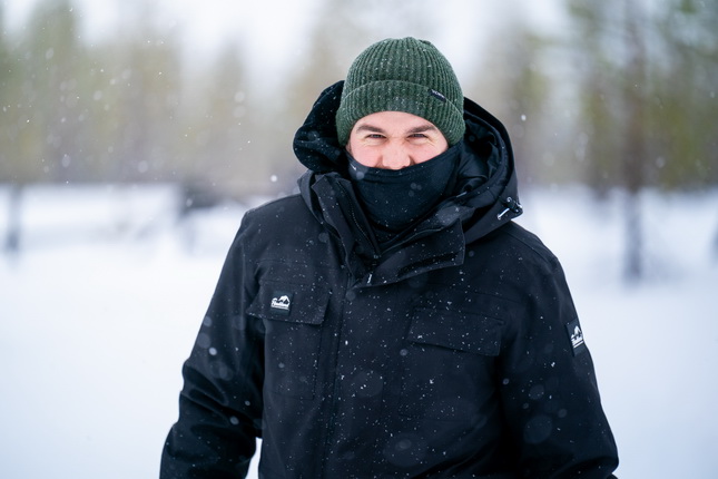 Ein Mann steht in der winterlichen Landschaft, die von fallendem Schnee bedeckt ist. Er trägt eine schwarze Winterjacke und einen grünen Mütze. Sein Gesicht ist teilweise von einem schwarzen Schal verdeckt, während er in die Kamera lächelt. Im Hintergrund sind verschwommene, schneebedeckte Bäume zu sehen.