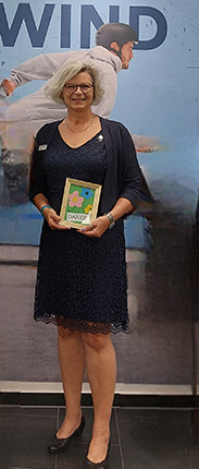 Eine Frau in einem eleganten, dunkelblauen Kleid steht in einer Ausstellung. Sie hält einen Bilderrahmen mit einem bunten Kunstwerk, das eine blühende Pflanze zeigt. Im Hintergrund ist ein großes Bild eines Mannes, der auf einem Skateboard fährt. Die Umgebung ist modern und professionell gestaltet.