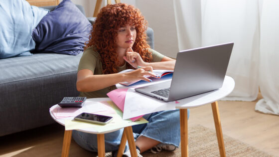 Eine Frau mit lockigem, rotem Haar sitzt auf dem Boden und arbeitet an einem Laptop, umgeben von Unterlagen und einem Taschenrechner. Sie trägt ein grünes T-Shirt und eine Jeans. Auf einem kleinen Tisch liegen farbige Blätter und ein Smartphone.