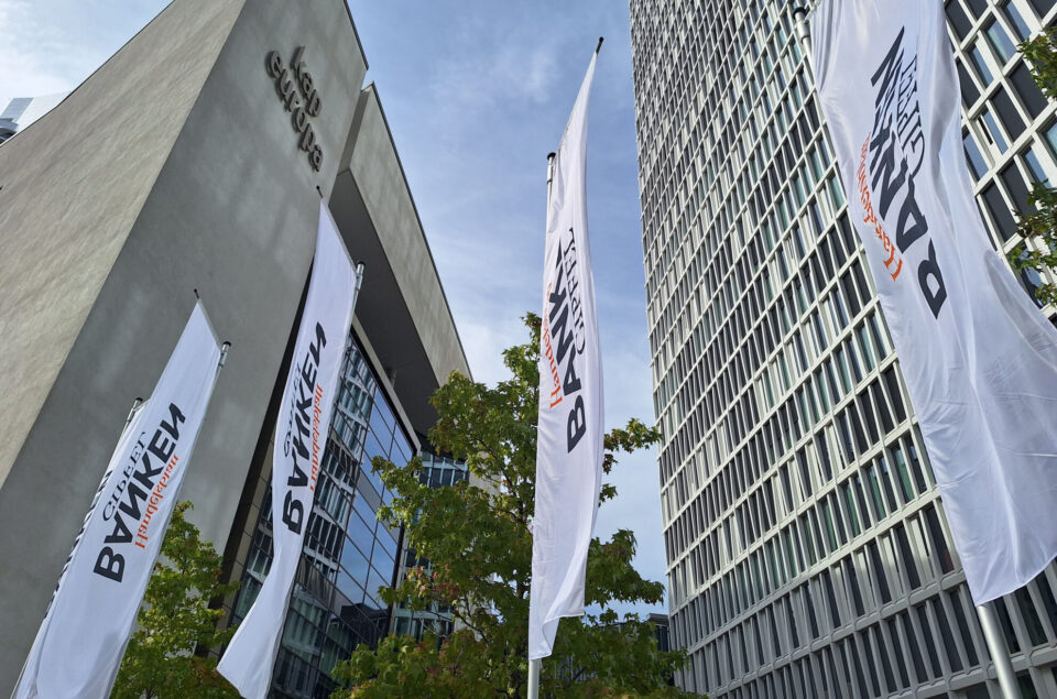 Die Aufnahme zeigt das Gebäude 'Kap Europa' mit grauer Fassade und modernem Design. Vor dem Gebäude stehen mehrere große Fahnen mit dem Schriftzug 'BAYKEN'. Im Hintergrund sind Glasfenster eines weiteren Gebäudes sichtbar. Der Himmel ist leicht bewölkt.