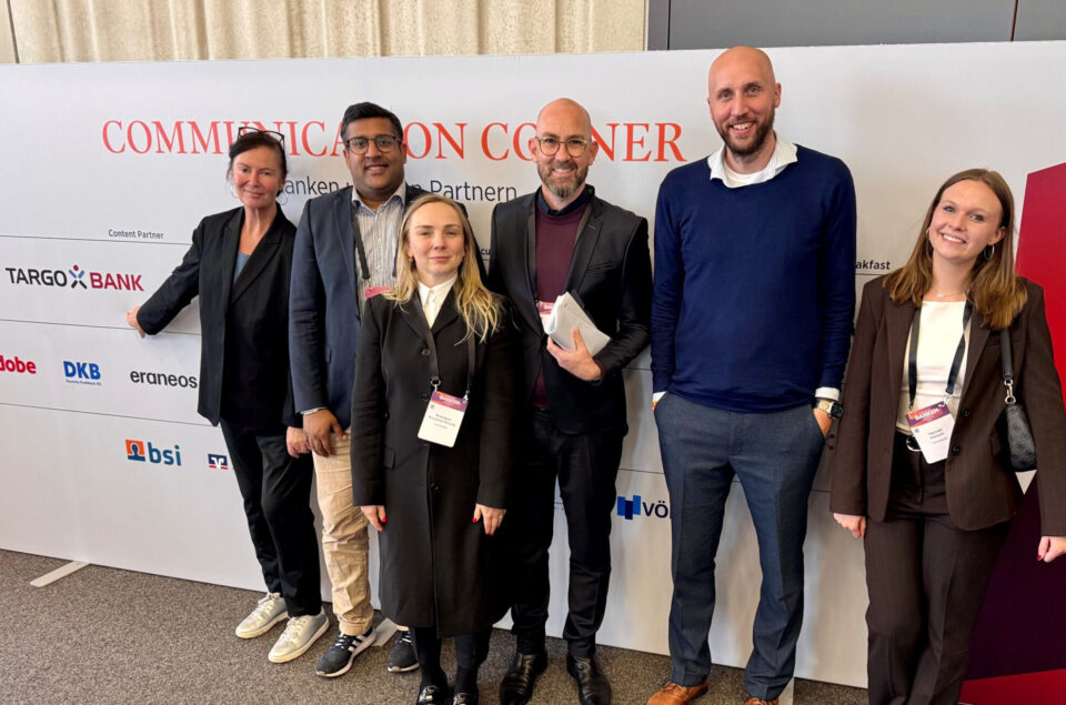 Sechs Personen stehen vor einem großen Schild mit der Aufschrift "COMMUNICATION CORNER". Sie sind in formeller Kleidung und lächeln. Auf dem Schild sind Logos verschiedener Partner wie TARGO BANK, DKB und andere sichtbar. Eine Person zeigt auf das TARGO BANK-Logo. Die Gruppe steht in einem hellen Raum mit einem Teppichboden.