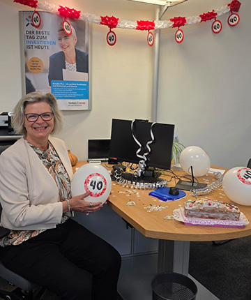Eine Frau sitzt an einem Schreibtisch mit einem Lächeln und hält einen Ballon mit der Zahl 40. Der Schreibtisch ist festlich dekoriert mit Luftballons, Konfetti und einer Torte. Im Hintergrund hängt eine Girlande mit der Zahl 40. Es gibt einen Computer und eine Plakatwand mit Text, der besagt: 'DER BESTE TAG ZUM INVESTIEREN IST HEUTE'.