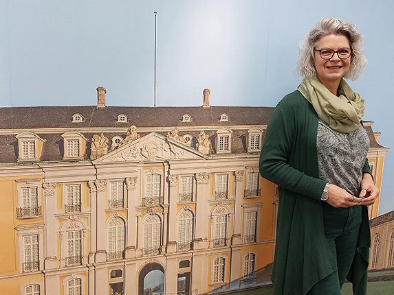 Eine Frau mit grauen Haaren und Brille steht vor einer großen Wandgrafik eines historischen Gebäudes. Sie trägt eine grüne Jacke und einen hellen Schal.