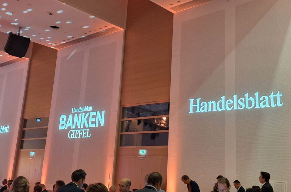 Ein Konferenzraum mit einer Veranstaltung namens "Handelsblatt Banken Gipfel". An den Wänden sind die Schriftzüge "Handelsblatt" und "BANKEN GIPFEL" projiziert. Der Raum hat eine elegante Atmosphäre mit sanftem Licht.