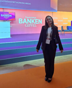 Eine Frau in einem schwarzen Anzug steht lächelnd auf einer orangefarbenen Bühne. Im Hintergrund sind Stühle und ein Bildschirm mit dem Text 'Handelsblatt BANKEN GIPFEL 2026' und 'anschließend NETWORKING-BREAK' sichtbar.