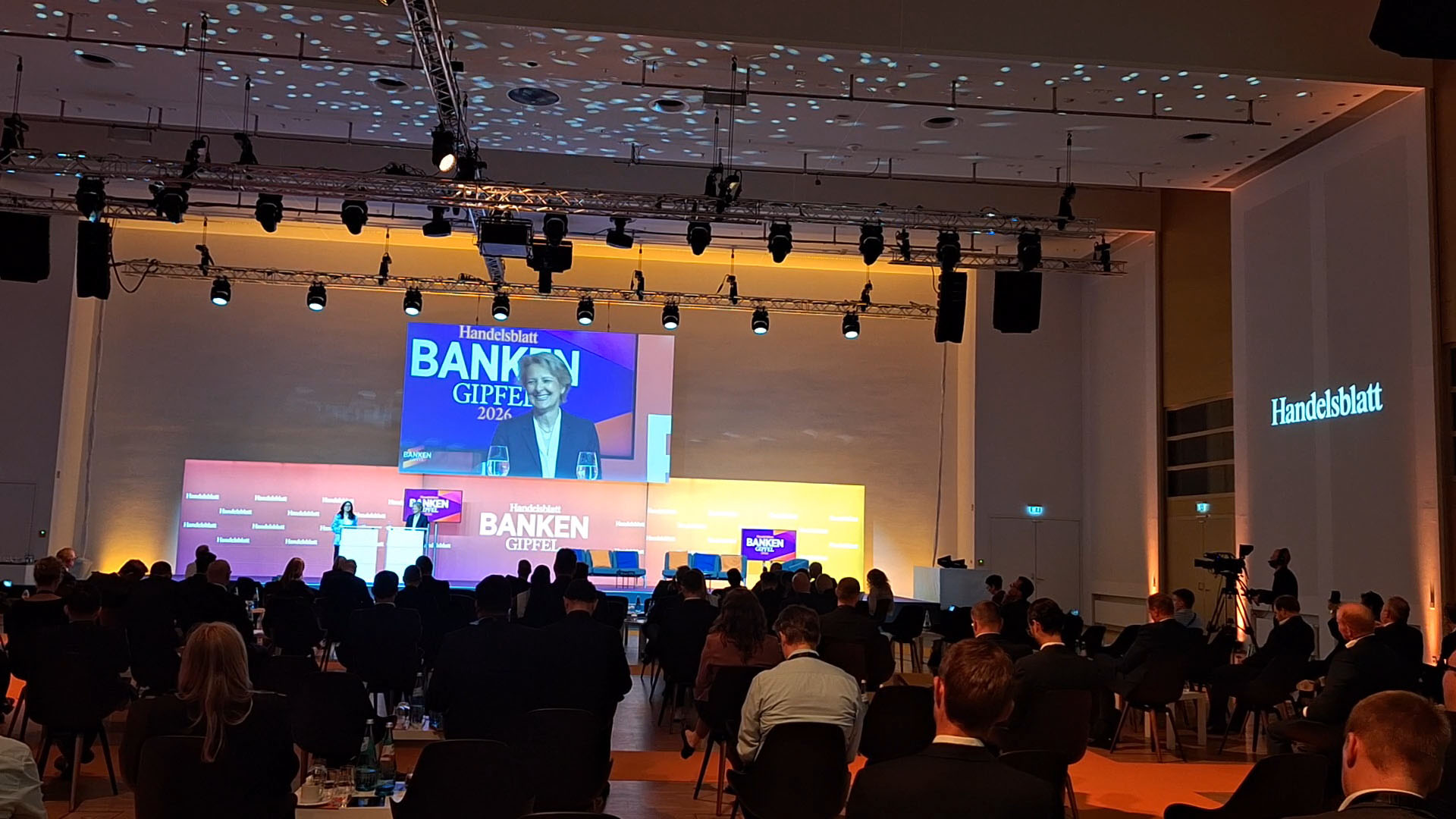 Eine Konferenz mit vielen Teilnehmern, die in einem großen Raum sitzen. Auf der Bühne steht eine Frau mit einem Mikrofon, und im Hintergrund ist ein großes Display mit dem Text 'Handelsblatt BANKEN GIPFEL 2023'. Die Atmosphäre ist professionell, mit Scheinwerfern und einer Bühne, die mit Stühlen ausgestattet ist. Es sind Getränke auf dem Podium sichtbar, und die Zuschauer scheinen aufmerksam zu sein.