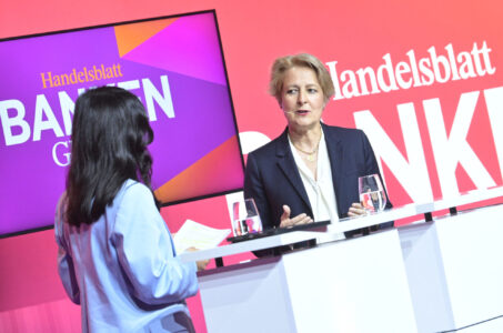 Eine Gesprächsrunde auf einer Konferenz. Eine Frau mit blonden Haaren spricht, während eine andere Frau in einem blauen Blazer zuhört. Im Hintergrund ist ein Bildschirm mit dem Schriftzug 'Handelsblatt Banken' zu sehen.