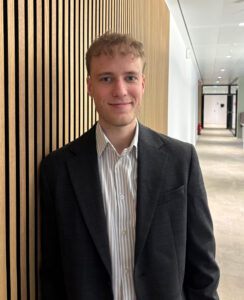 Ein junger Mann mit lockigem Haar trägt ein schwarzes Sakko über einem gestreiften Hemd. Er steht an einer holzverkleideten Wand in einem modernen Bürogebäude, mit einem Flur im Hintergrund.