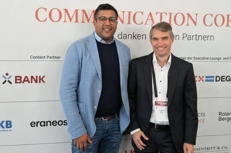 Zwei Männer stehen lächelnd vor einer Wand mit dem Schriftzug 'COMMUNICATION CORP'. Der Mann links trägt einen hellblauen Blazer, ein dunkelblaues Hemd und eine Brille. Der Mann rechts hat graue Haare, trägt einen schwarzen Anzug und ein weißes Hemd. Beide scheinen an einer Veranstaltung teilzunehmen, die Sponsorenlogos im Hintergrund zeigt.