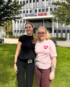 Zwei Frauen stehen auf einer grünen Wiese vor einem modernen Gebäude mit dem Schriftzug 'TARGOBANK'. Die Frau links trägt ein schwarzes Oberteil und hat lange Haare. Die Frau rechts trägt ein rosa T-Shirt mit einem Herz und hat blonde, lockige Haare. Beide lächeln in die Kamera.