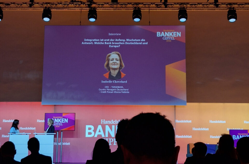 Auf der Bühne eines Events sitzen zwei Personen. Ein Bildschirm zeigt das Interview mit Isabelle Chevelard, CEO der TARGOBANK. Der Text beschreibt die Themen Integration und Wachstum in Bezug auf Banken in Deutschland und Europa.