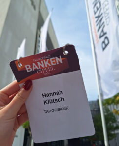 Eine Hand hält ein Namensschild mit dem Text 'Hannah Klütsch, TARGOBANK' und dem Titel 'Handelsblatt BANKEN GIPFEL 2026'. Im Hintergrund sind Fahnen mit dem Schriftzug 'BANKEN' und ein Gebäude sichtbar.