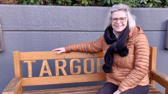 Eine Frau mit langen, grauen Haaren sitzt auf einer Holzbank mit der Aufschrift "TARGOB". Sie trägt eine braune, gepolsterte Jacke und einen schwarzen Schal. Im Hintergrund sind grüne Pflanzen zu sehen.