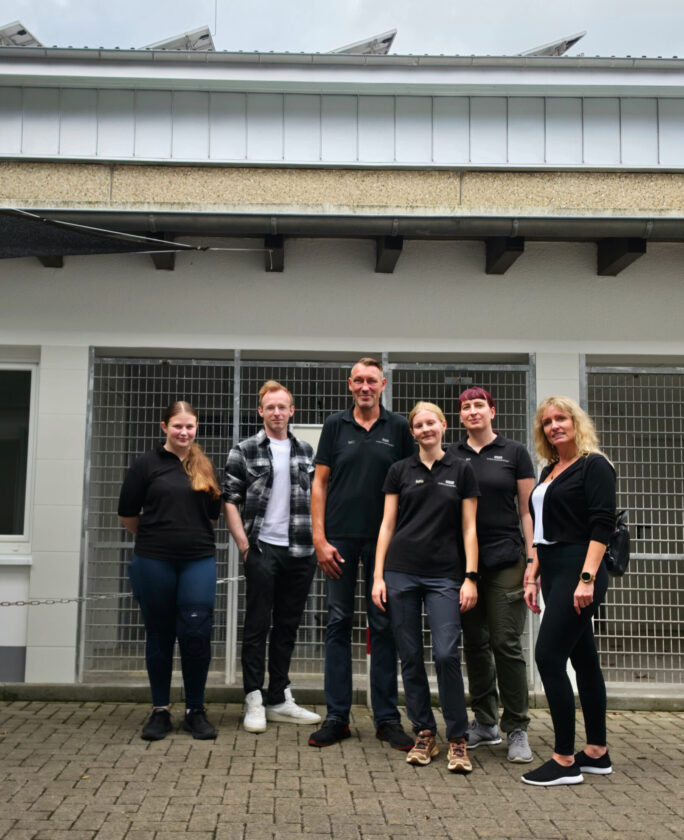 Sechs Personen stehen in einer Reihe vor einem Gebäude mit Fenster und einem Gitter. Die Gruppe besteht aus drei Frauen und drei Männern, die schwarze Poloshirts und andere legere Kleidung tragen. Die Männer stehen in der Mitte, ein Mann hat ein kariertes Hemd an. Der Hintergrund zeigt eine graue Wand und eine geklärte Struktur. Die Umgebung scheint urban und modern zu sein.