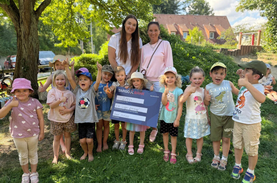 Eine Gruppe von Kindern und zwei Erwachsenen steht unter einem Baum. Die Kinder halten einen großen Scheck mit der Aufschrift "TARGOBANK" und "2000€". Alle lächeln und zeigen den Daumen hoch. Im Hintergrund sind einige Spielgeräte und ein Auto sichtbar.