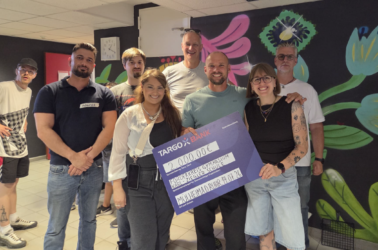 Eine Gruppe von Personen steht in einem Raum mit bunten Wänden. Sie halten einen großen Scheck von TARGO-Bank über 2000 €. Die Gruppe besteht aus Männern und Frauen, die lächeln und fröhlich aussehen.