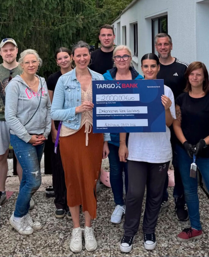 Eine Gruppe von Menschen, die vor einem Gebäude steht, hält einen großen Scheck der TARGOBANK über 2.000 Euro. Die Menschen lächeln und posieren für das Foto. Im Hintergrund sind Bäume und ein Teil des Gebäudes sichtbar.