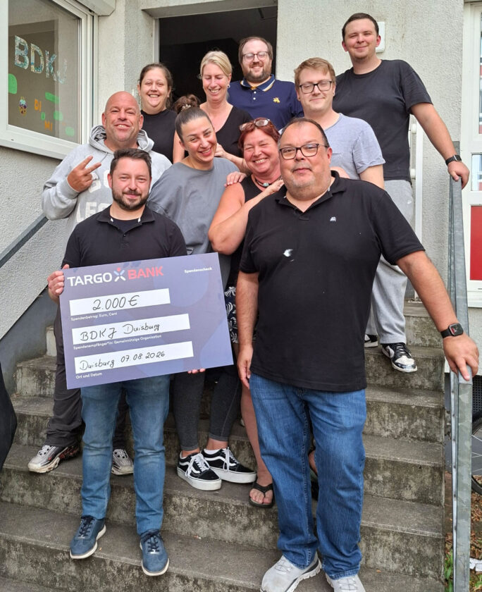 Eine Gruppe von zehn Personen steht auf einer Treppe vor einem Gebäude. Sie halten einen großen Scheck von TARGOBANK über 2.000 € für BDK Duisburg. Die Stimmung scheint erfreulich zu sein, einige lächeln und posieren für das Foto.