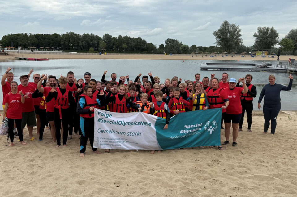 Eine große Gruppe von Menschen in Schwimmwesten posiert am Strand. Sie halten ein Banner mit dem Text 'Folge #SpecialOlympicsNRW Gemeinsam stark!' und 'Special Olympics Nordrhein-Westfalen'. Im Hintergrund ist ein ruhiger See und einige Bäume sichtbar.